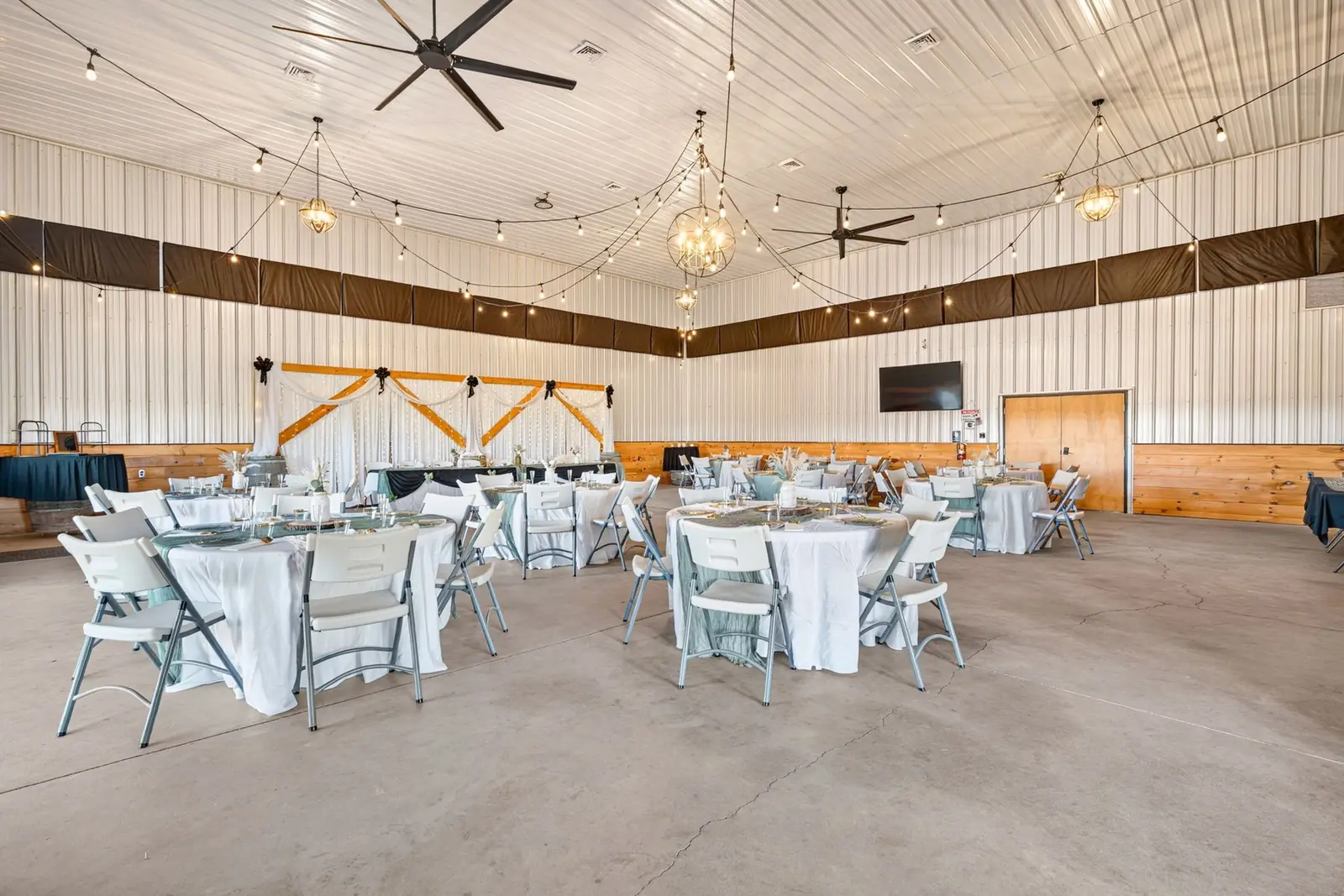 Reception hall at Barn at the Lucky Farm set for a wedding with round tables in white linens and sage runners, white chairs, globe chandeliers, string lights and a head table with a wood backdrop